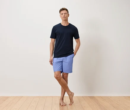 Un homme portant un t-shirt bleu marine et un short bleu clair se tient devant un mur blanc.