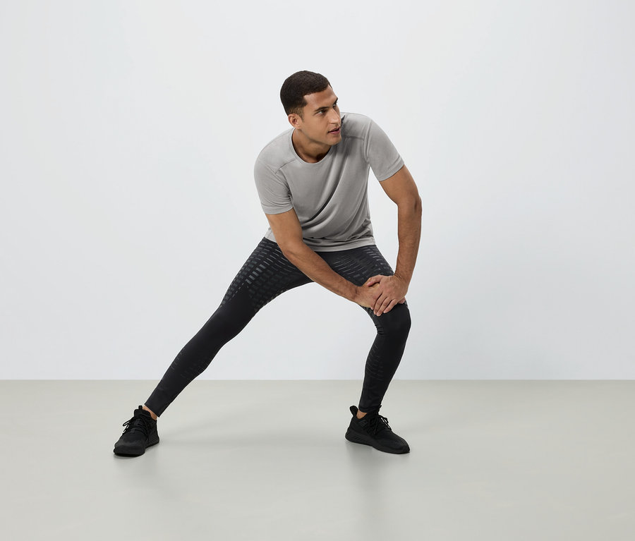 Un homme s'étire en t-shirt gris, legging noir et baskets noires sur fond blanc.