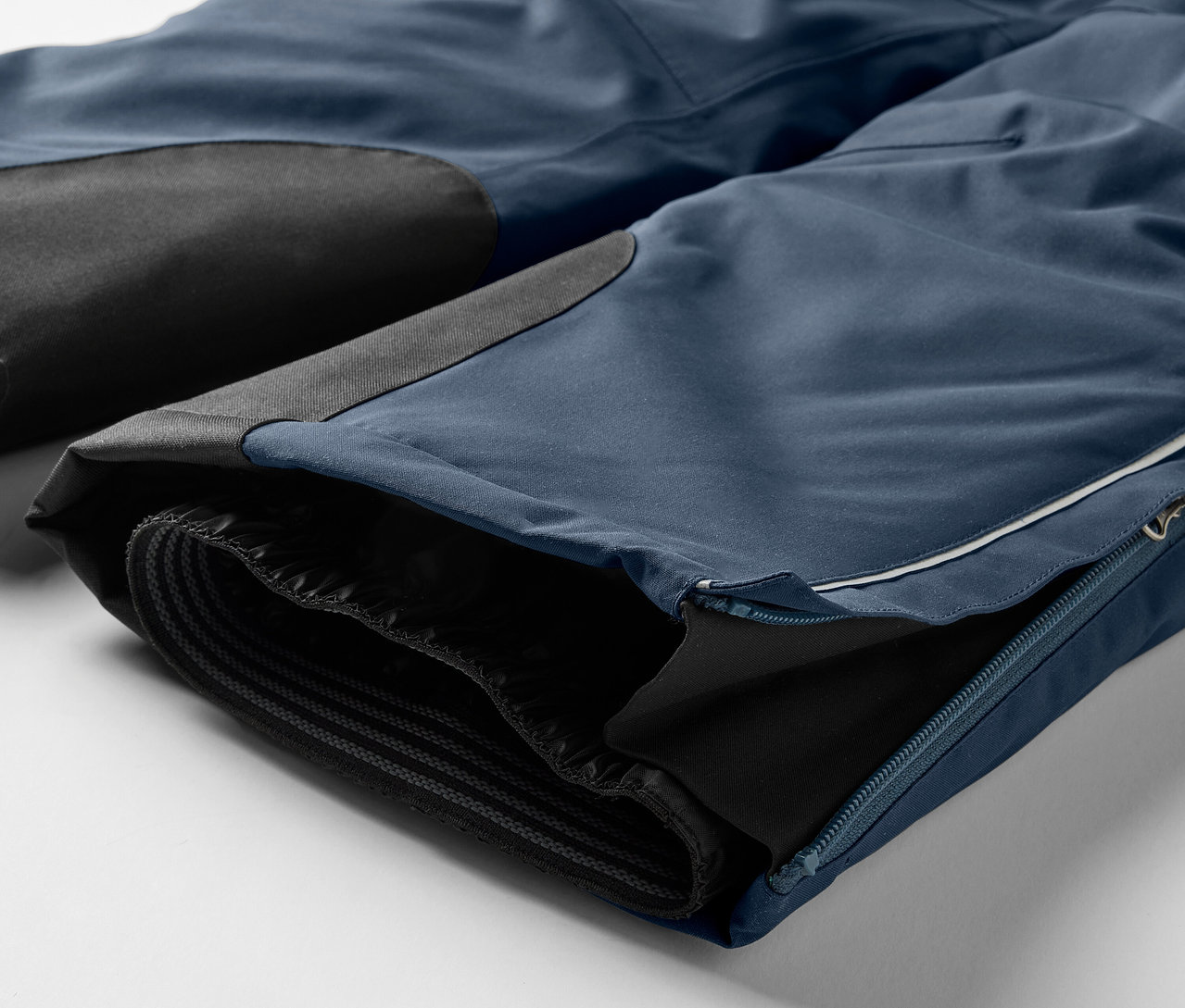 Détail d'un pantalon bleu avec des empiècements noirs aux genoux et une fermeture éclair sur la jambe.