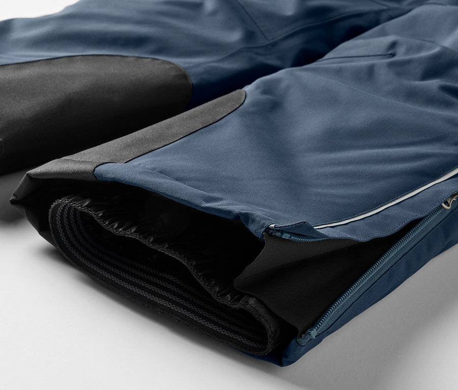 Détail d'un pantalon bleu avec des empiècements noirs aux genoux et une fermeture éclair sur la jambe.