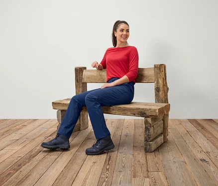 Une femme est assise sur un banc en bois, portant un chemisier rouge, un pantalon bleu et des bottes.
