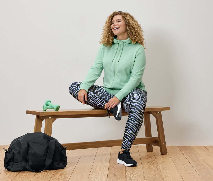 Une femme aux cheveux bouclés est assise sur un banc en bois, portant un sweat à capuche vert menthe et un legging à motif zébré. Un sac de sport noir et des haltères vertes sont également visibles.