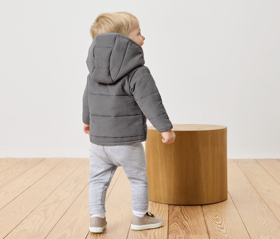 Un petit garçon blond porte une veste matelassée grise à capuche, un pantalon gris et des chaussures beiges. Il se tient sur un plancher en bois à côté d'un tabouret rond en bois.