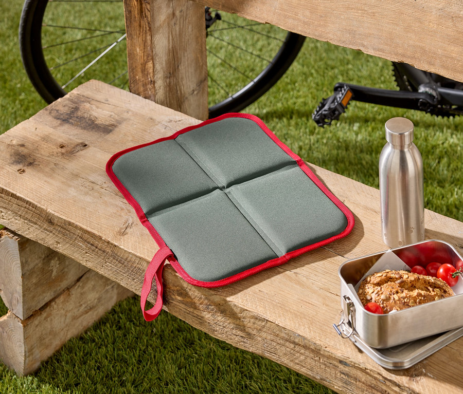 Un coussin de siège pliable gris avec un bord rouge est posé sur un banc en bois, à côté d'une boîte à pain et d'une bouteille en acier inoxydable. Un vélo est visible à l'arrière-plan.