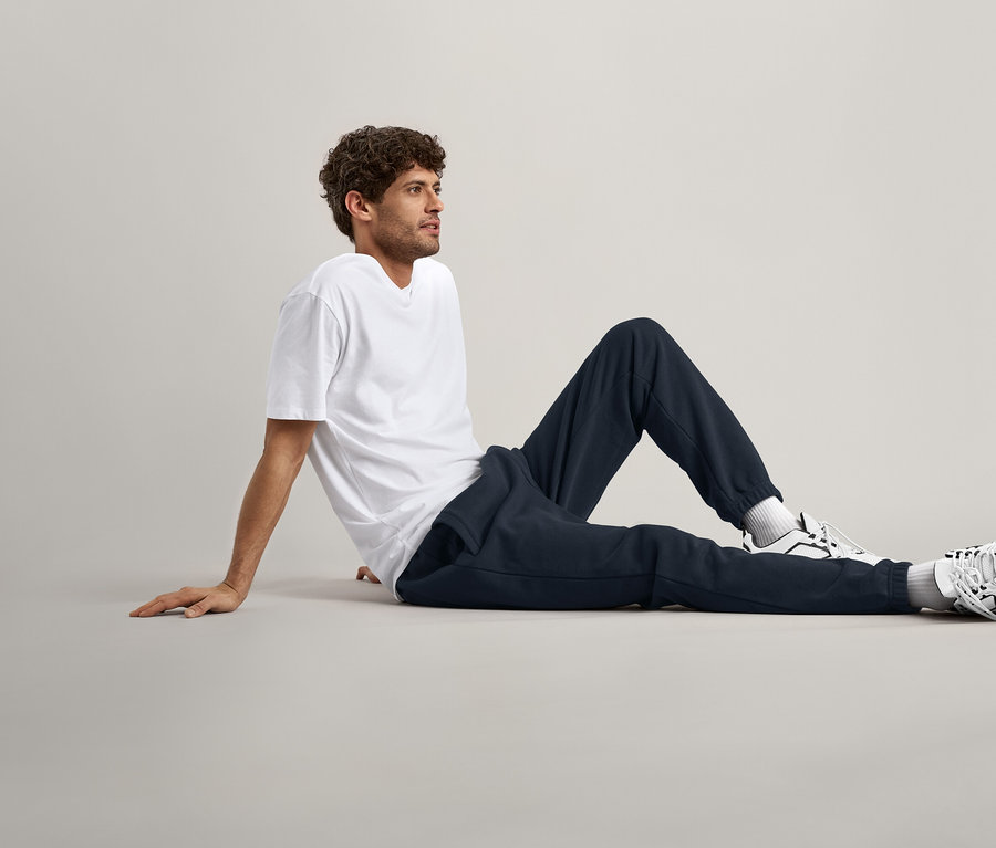 Homme aux cheveux bouclés assis par terre, portant un t-shirt blanc, un pantalon de survêtement bleu marine et des baskets.