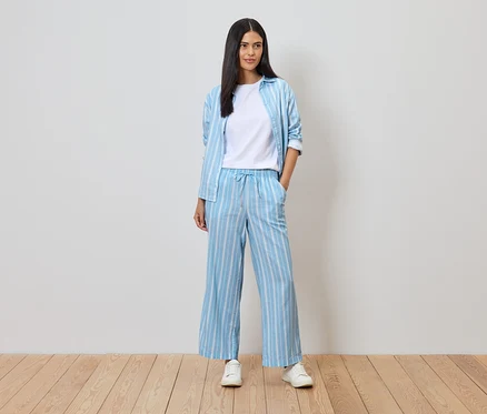 Femme portant un pantalon et une chemise bleu et blanc rayés sur un t-shirt blanc, avec des baskets blanches sur un plancher en bois devant un mur blanc.