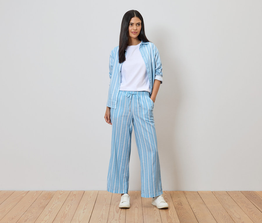 Femme portant un pantalon et une chemise bleu et blanc rayés sur un t-shirt blanc, avec des baskets blanches sur un plancher en bois devant un mur blanc.