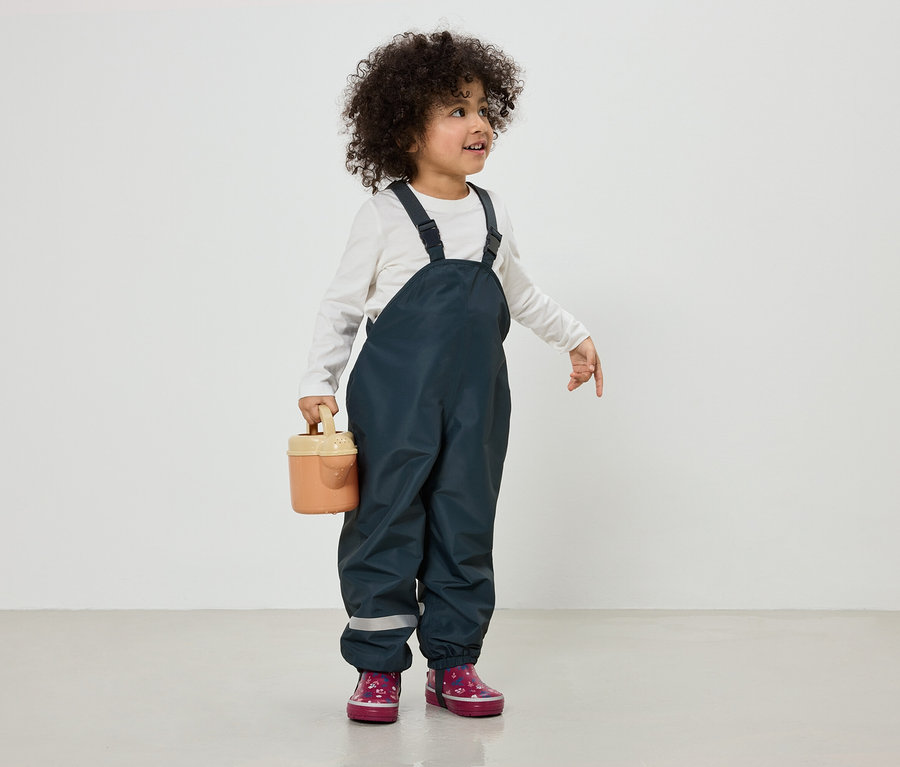 Un enfant aux cheveux bouclés porte une salopette bleu marine et des bottes roses, et tient un petit arrosoir.