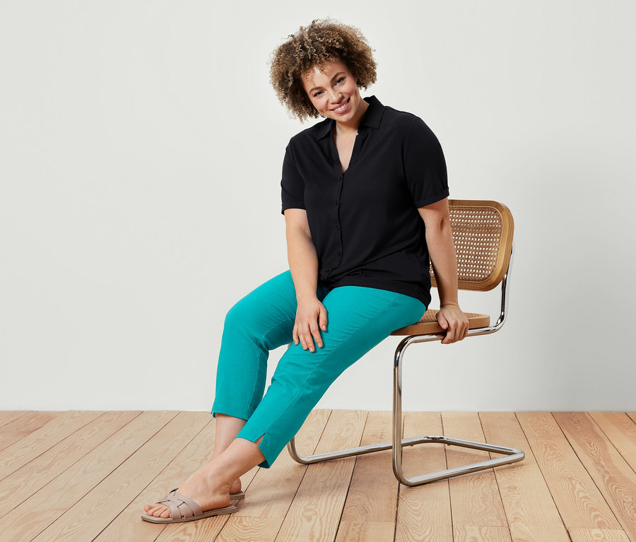 Une femme assise sur une chaise porte un chemisier noir et un pantalon stretch 7/8 bleu azur.