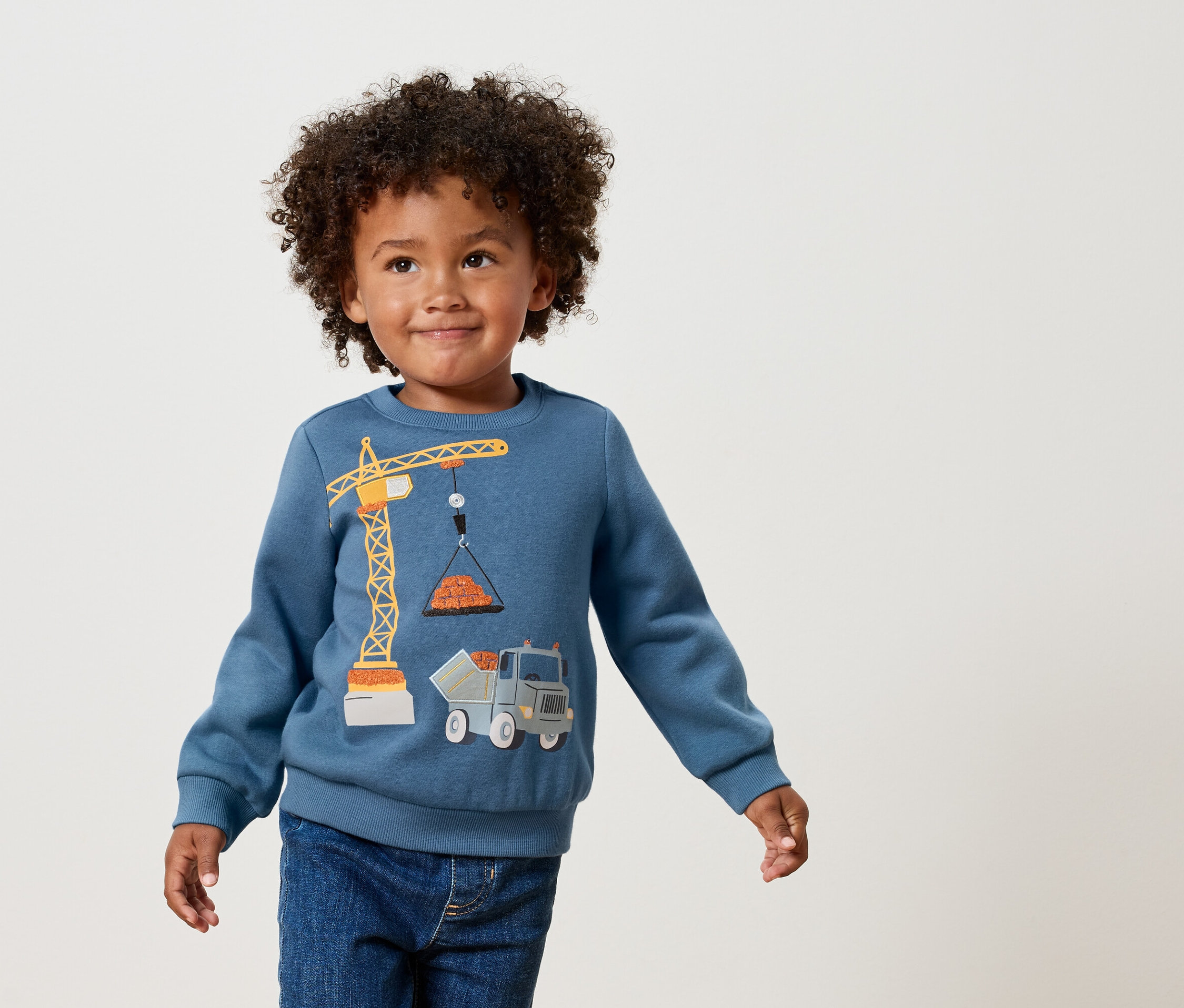Un garçon aux cheveux bouclés porte un sweat-shirt bleu avec une application de grue et de camion jaune.