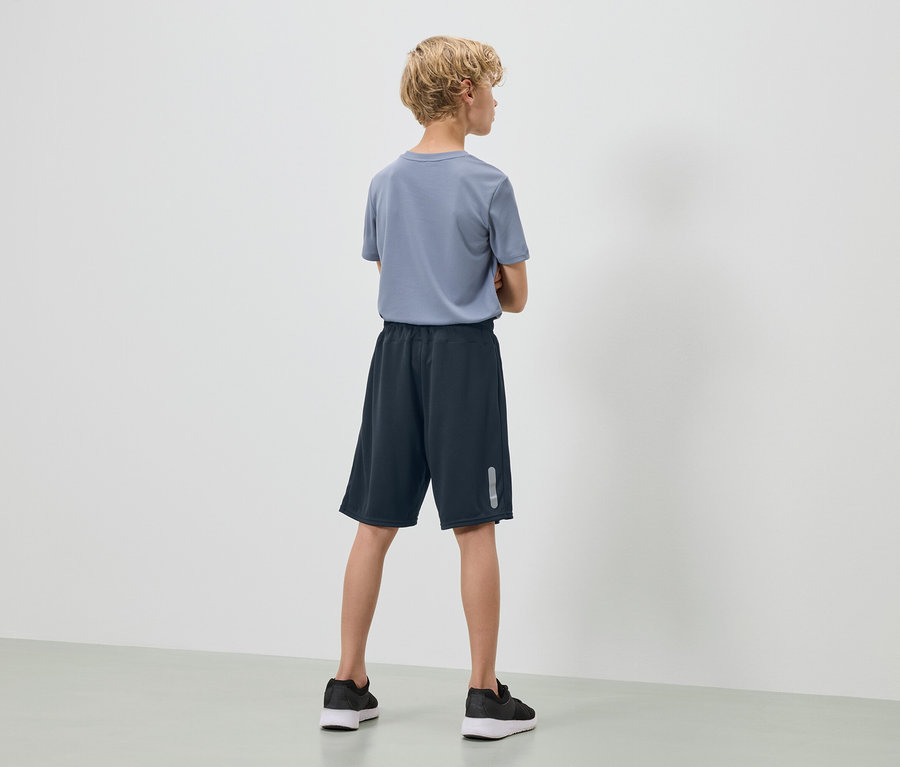 Un adolescent aux cheveux blonds et bouclés se tient dos à la caméra, portant un t-shirt bleu, un short bleu marine et des baskets noires.