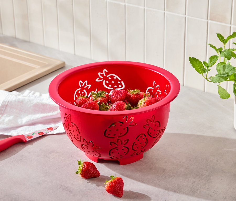 Une passoire rouge à motif de fraises remplie de fraises, à côté de laquelle se trouve un couteau à motif de fraises.