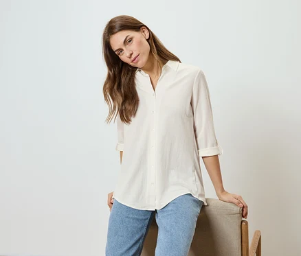 Femme aux cheveux ondulés, portant une chemise claire et un jean, appuyée sur une chaise.
