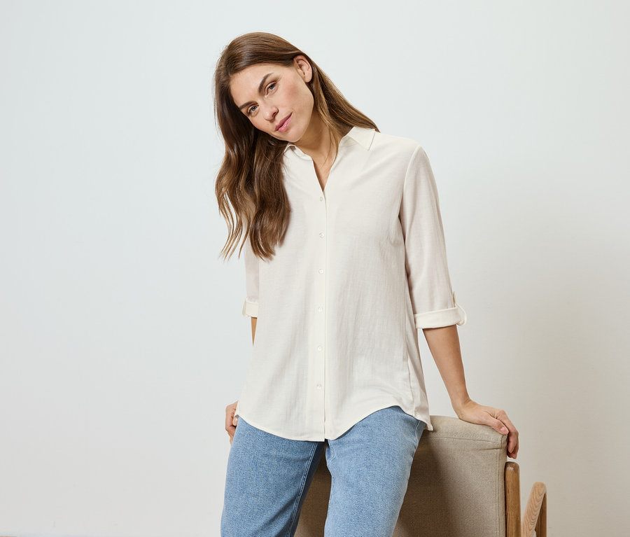 Femme aux cheveux ondulés, portant une chemise claire et un jean, appuyée sur une chaise.