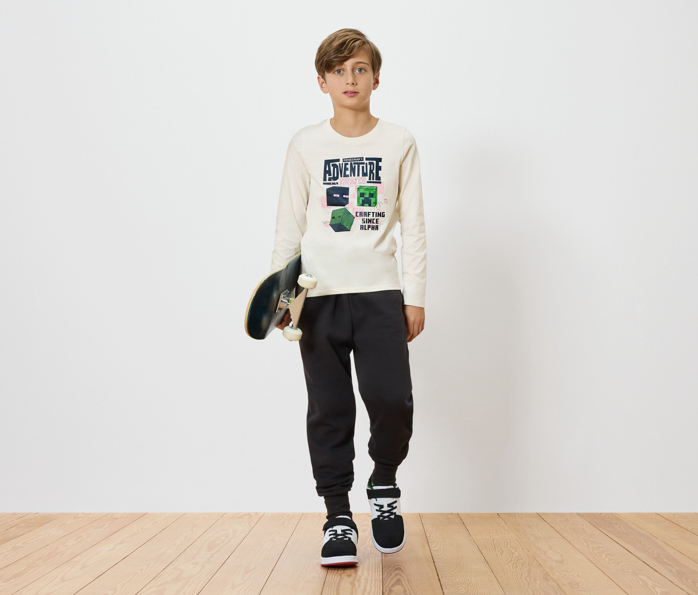 Un garçon tient un skateboard et porte un t-shirt à manches longues clair avec un motif Minecraft, associé à un pantalon de jogging foncé et des baskets sur un fond uni.