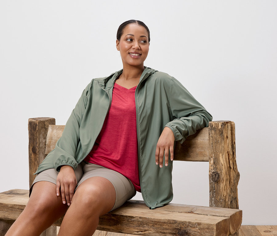 Femme assise sur un banc en bois portant une veste verte, une chemise rouge et un short.