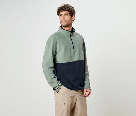 Un homme aux cheveux bouclés porte un haut en polaire bicolore vert et marine avec un short beige.