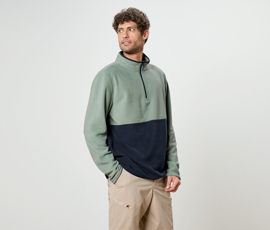 Un homme aux cheveux bouclés porte un haut en polaire bicolore vert et marine avec un short beige.