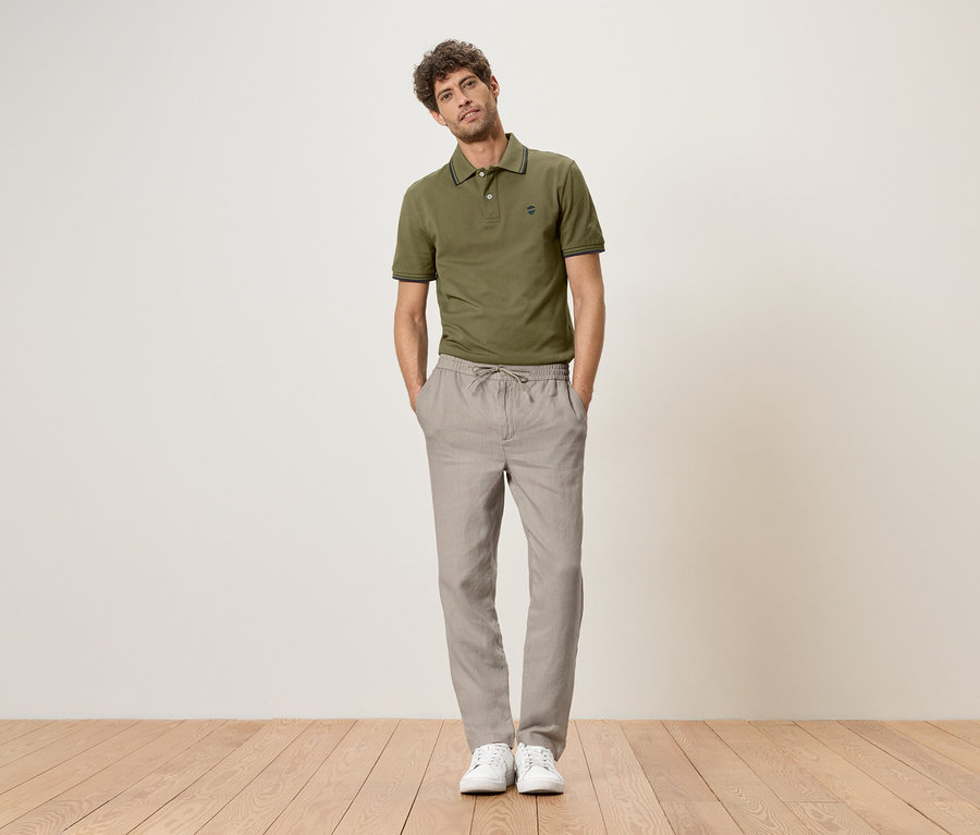 Un homme se tient debout, les mains dans les poches, portant un polo vert, un pantalon de lin gris et des baskets blanches.