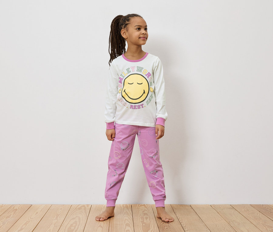 Une fillette avec des tresses se tient pieds nus dans un t-shirt blanc à manches longues avec un smiley et un pantalon rose avec des smileys.