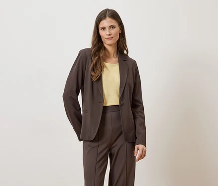 Femme portant un blazer et un pantalon marron avec un haut jaune sur fond clair.