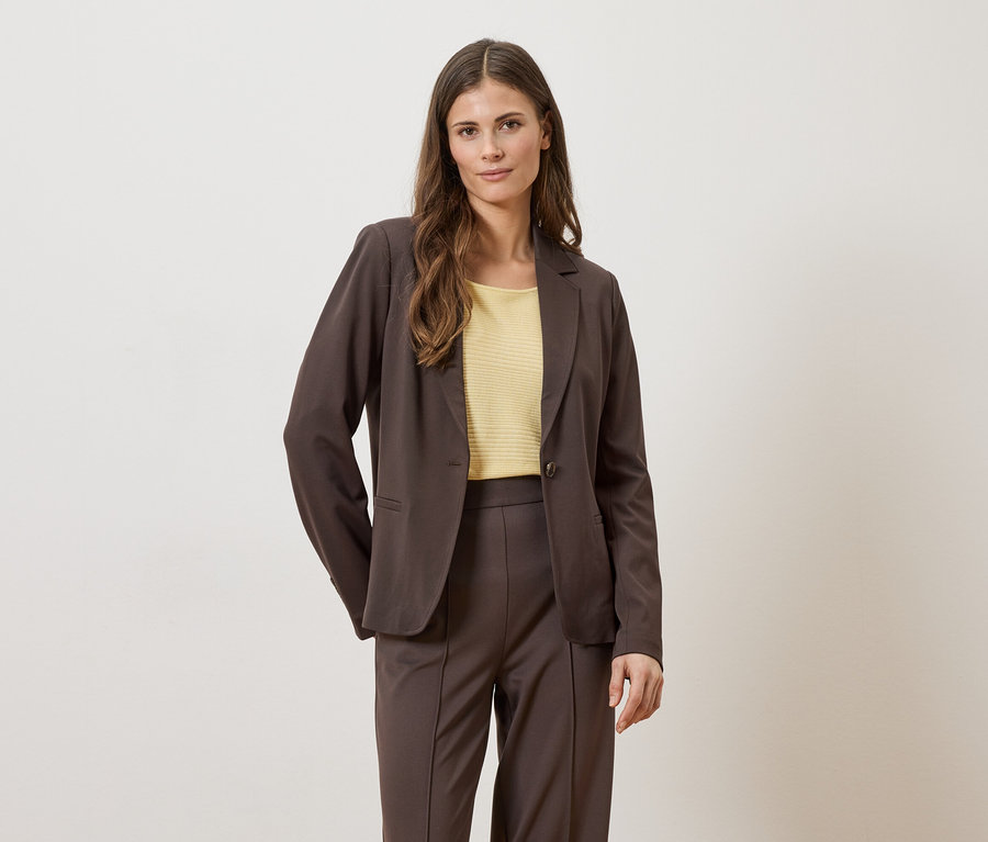 Femme portant un blazer et un pantalon marron avec un haut jaune sur fond clair.
