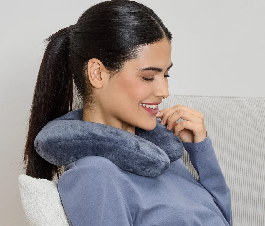 Une femme souriante, les cheveux attachés en queue de cheval et portant un haut bleu, a un coussin de voyage gris autour du cou.