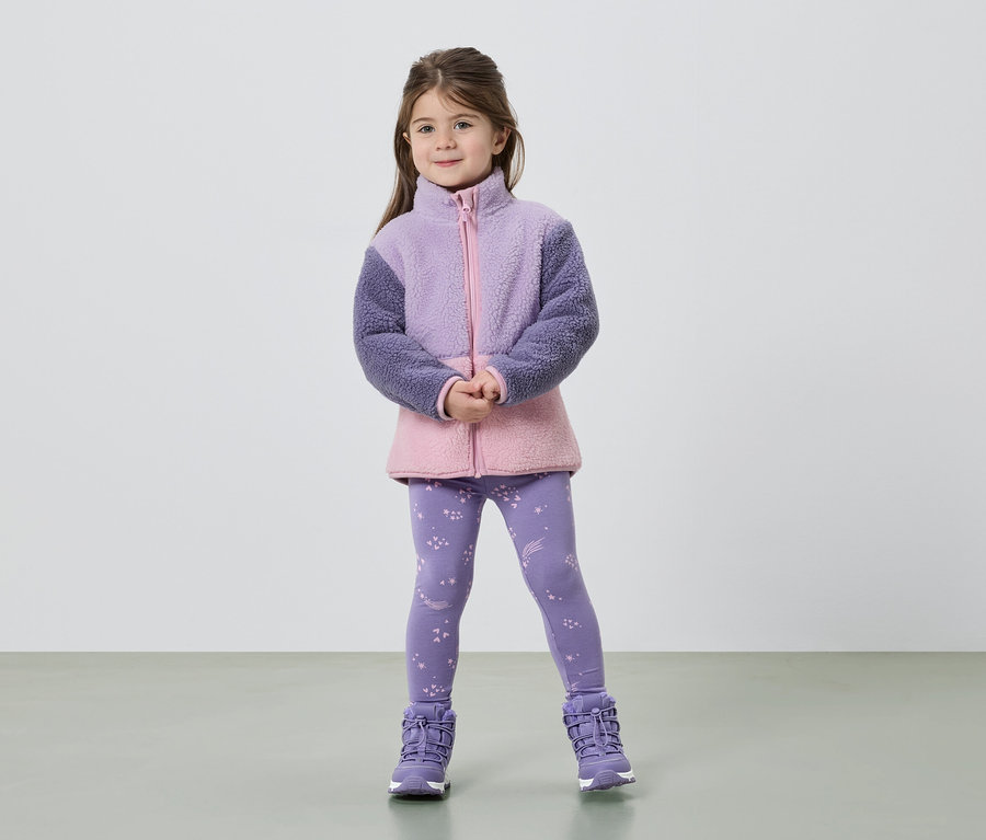 Une fillette se tient debout, les mains croisées, portant une veste en polaire violette et rose, un legging à étoiles et des bottes violettes.