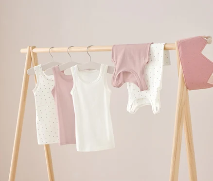 Sur un portant en bois sont suspendus trois débardeurs en blanc, rose et blanc à pois, ainsi qu'une chemise rose et un body blanc à pois. À droite est suspendue une couronne rose.