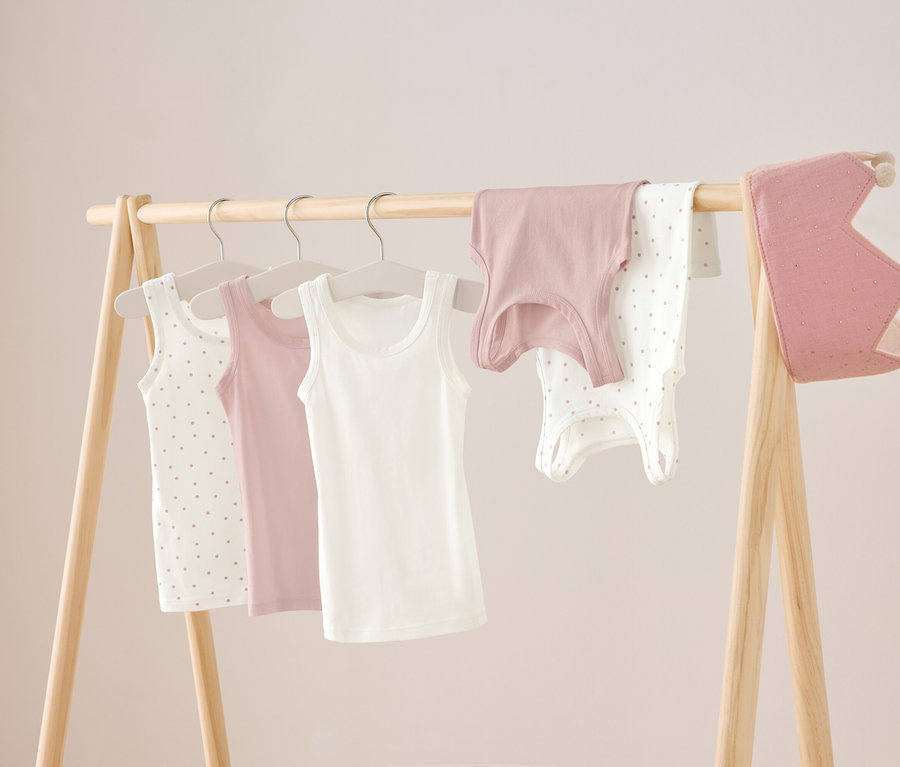 Sur un portant en bois sont suspendus trois débardeurs en blanc, rose et blanc à pois, ainsi qu'une chemise rose et un body blanc à pois. À droite est suspendue une couronne rose.