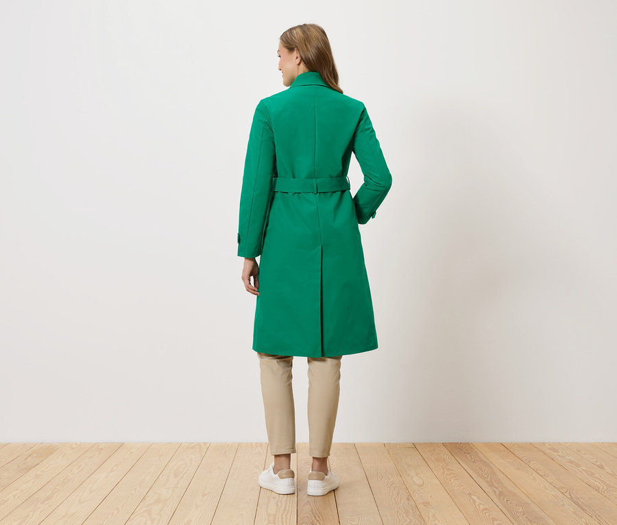Une femme se tient dos à la caméra et porte un manteau vert avec une ceinture, un pantalon beige et des baskets blanches.