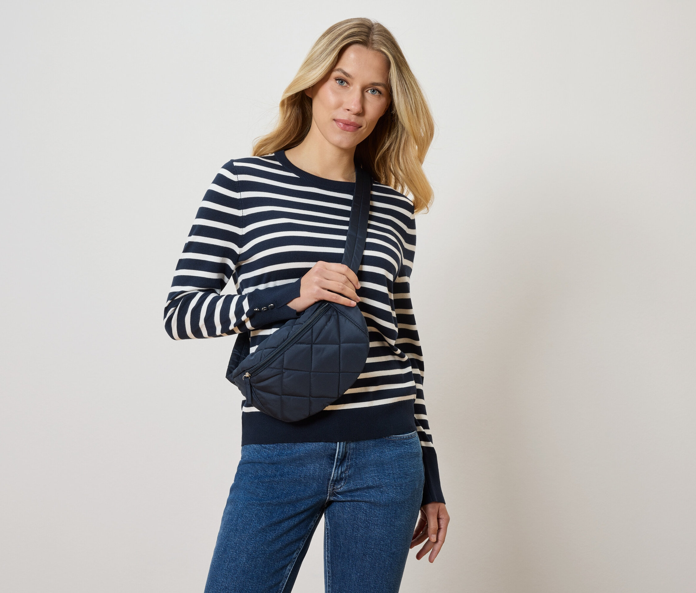 Femme blonde portant un pull rayé bleu et blanc et un jean bleu, tenant un sac banane marine.