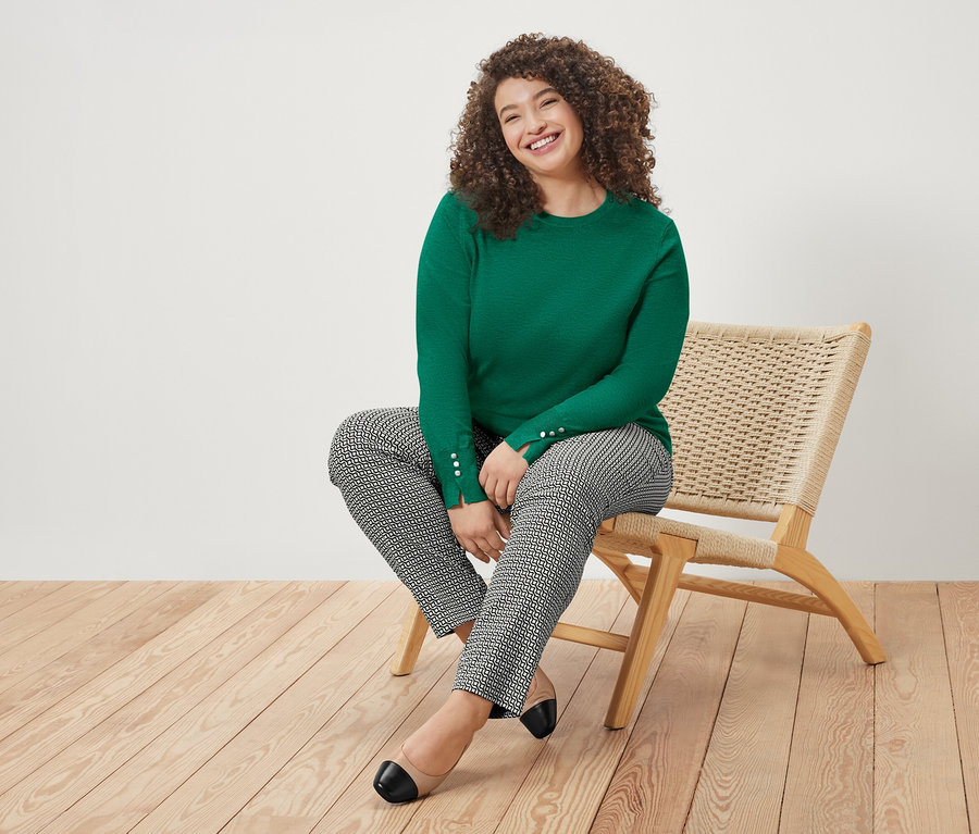 Femme aux cheveux bouclés assise sur une chaise, portant un pull vert et un pantalon à motifs.