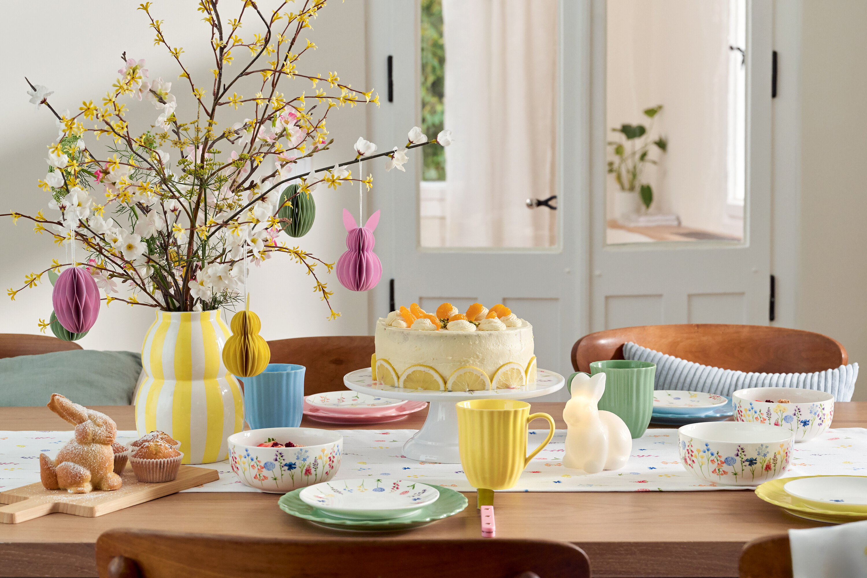 Table à manger avec décorations de Pâques, gâteau, vaisselle et vase de fleurs.