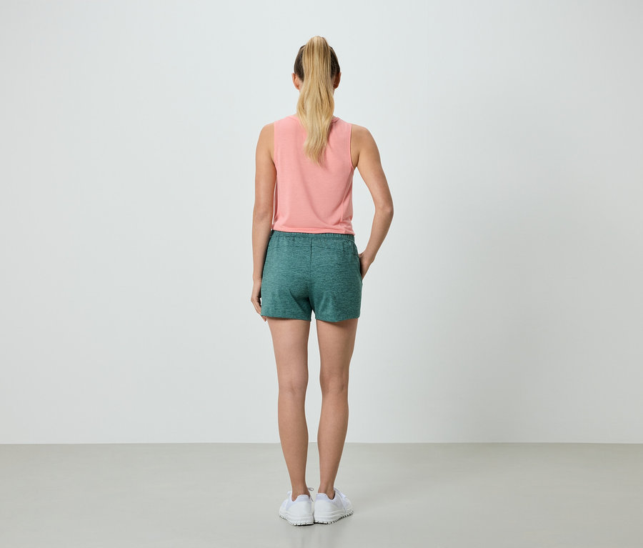Vue arrière d'une femme blonde portant un débardeur saumon et un short vert, debout en baskets blanches sur un fond neutre.