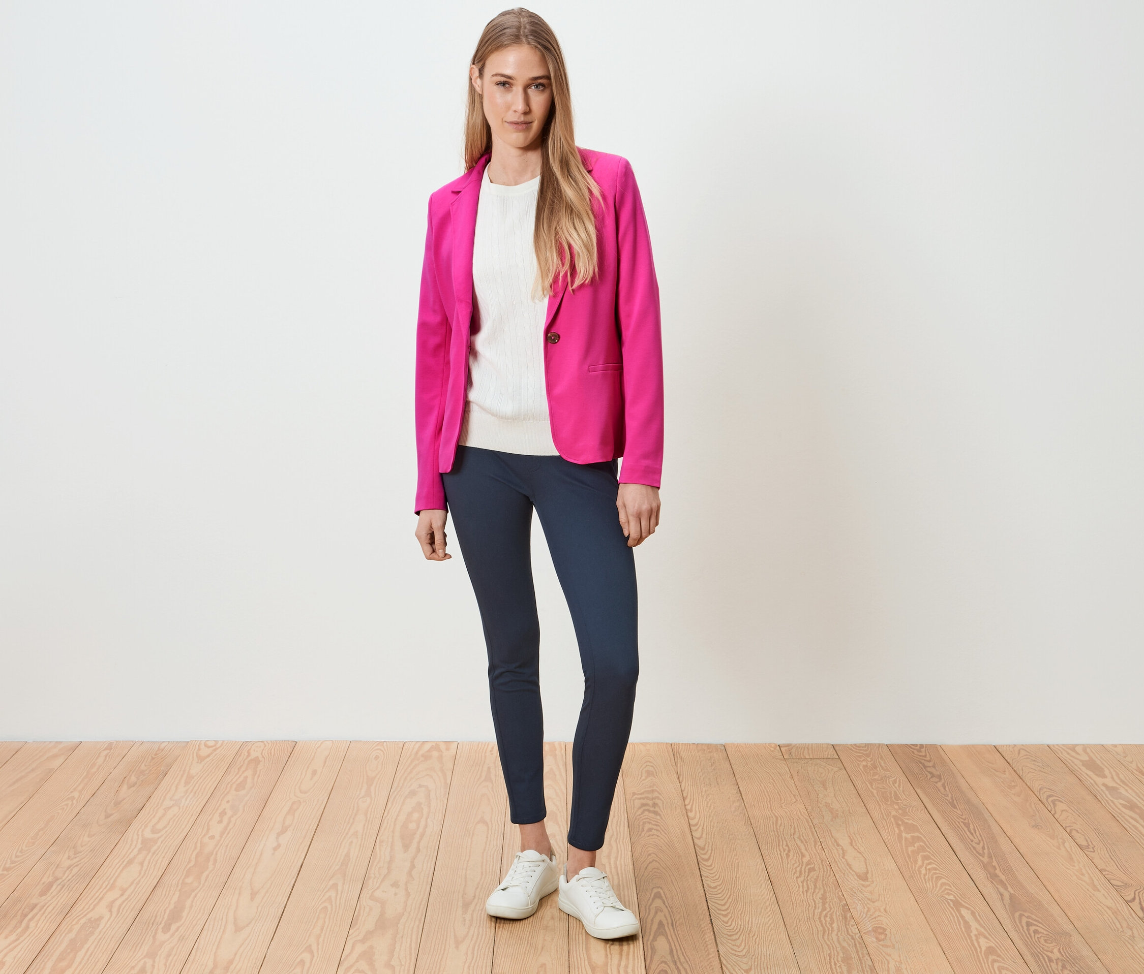 Femme debout portant un blazer rose, un haut blanc, un legging bleu et des baskets blanches.