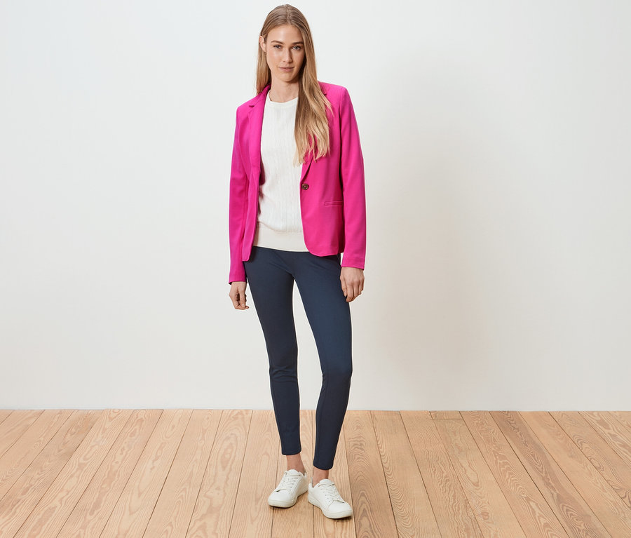 Femme debout portant un blazer rose, un haut blanc, un legging bleu et des baskets blanches.