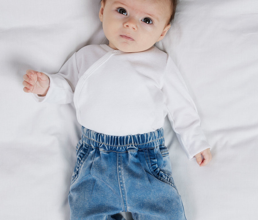 Vue de dessus d'un bébé portant un body cache-cœur blanc et un jean bleu clair sur un drap blanc.