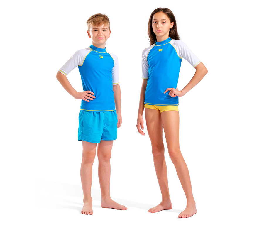 Un garçon et une fille se tiennent pieds nus devant un fond blanc. Ils portent tous les deux des hauts bleus et blancs et des maillots de bain.