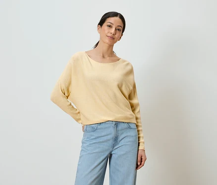 Une femme portant un pull jaune et un jean bleu pose devant un fond blanc.
