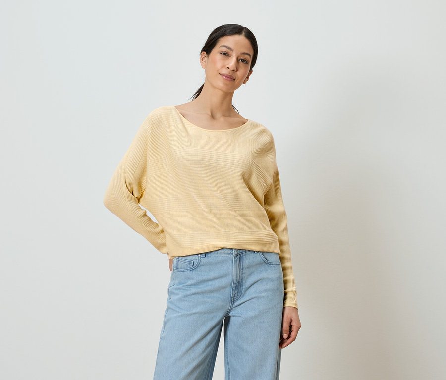 Une femme portant un pull jaune et un jean bleu pose devant un fond blanc.
