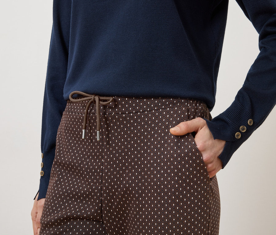 Personne portant un pull bleu marine et un pantalon marron à motifs avec cordon de serrage et main dans la poche.