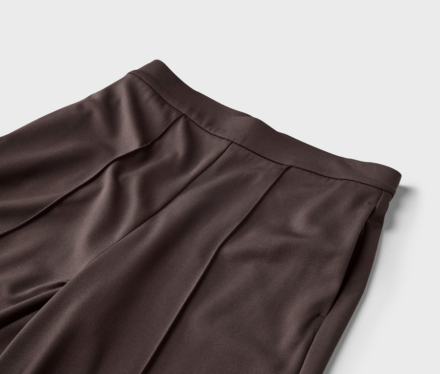 Gros plan de la taille d'un pantalon marron foncé avec des plis et une poche latérale sur un fond gris clair.