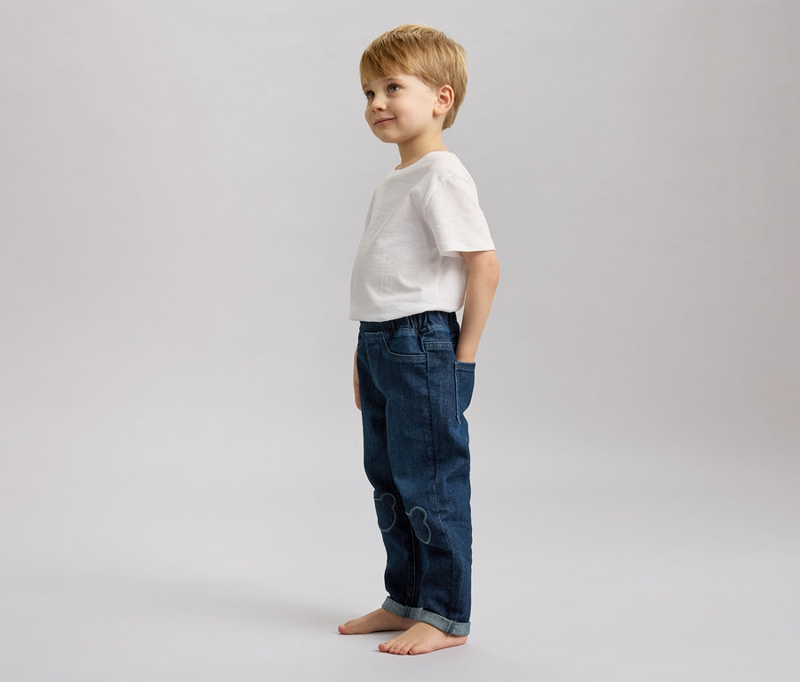 Un garçon blond se tient pieds nus en jean et t-shirt blanc, regardant vers la gauche.