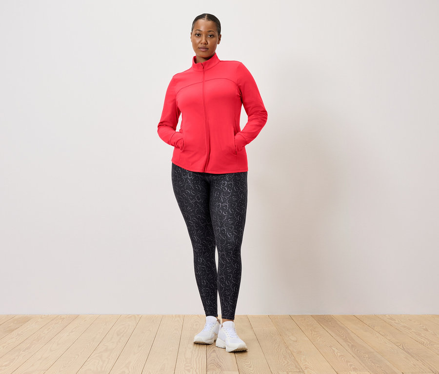 Femme portant une veste rouge et un legging à motifs posant devant un mur.