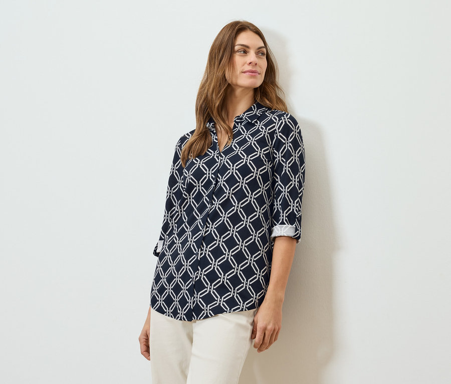 Femme aux cheveux bruns portant un chemisier bleu marine à motifs géométriques et un pantalon crème.