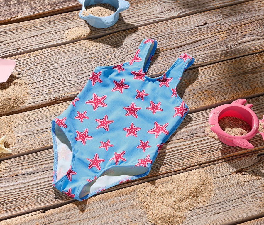 Un maillot de bain bleu clair à motifs d'étoiles de mer rouges est posé sur une surface en bois, avec des jouets de plage et du sable.