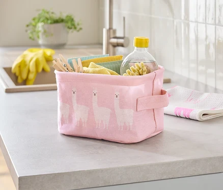 Sur un comptoir de cuisine se trouve une boîte en tissu rose à motif de lama, remplie de chiffons de nettoyage, de pinces à linge et d'une bouteille de nettoyant.