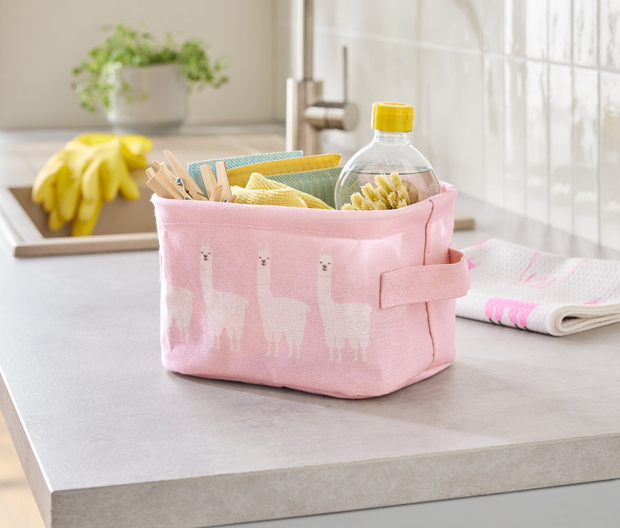 Sur un comptoir de cuisine se trouve une boîte en tissu rose à motif de lama, remplie de chiffons de nettoyage, de pinces à linge et d'une bouteille de nettoyant.