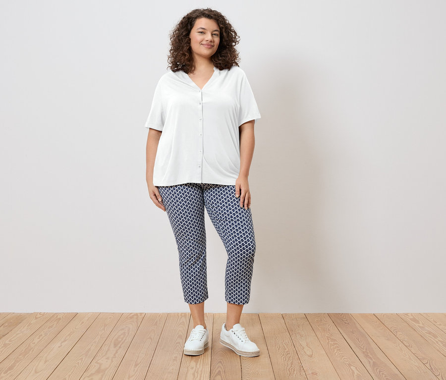 Une femme aux cheveux bouclés porte une chemise blanche et un pantalon à motifs bleus, ainsi que des chaussures blanches.